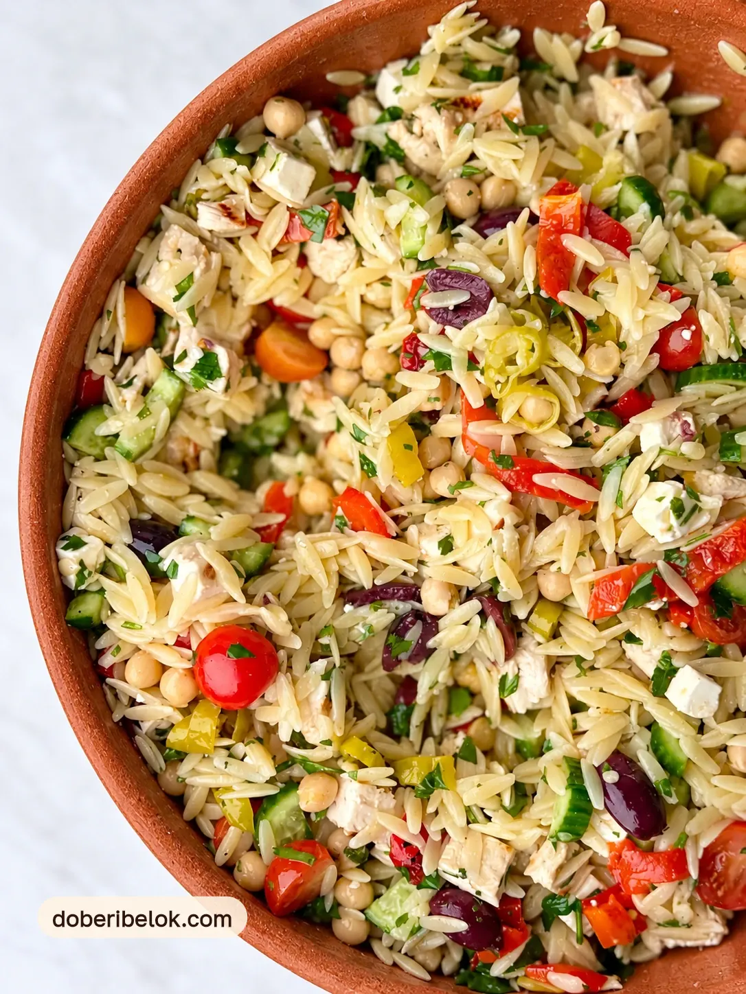 Салат из орзо с нутом, курицей, фетой, огурцами, черри, маслинами, перцем и петрушкой крупным планом в терракотовой миске